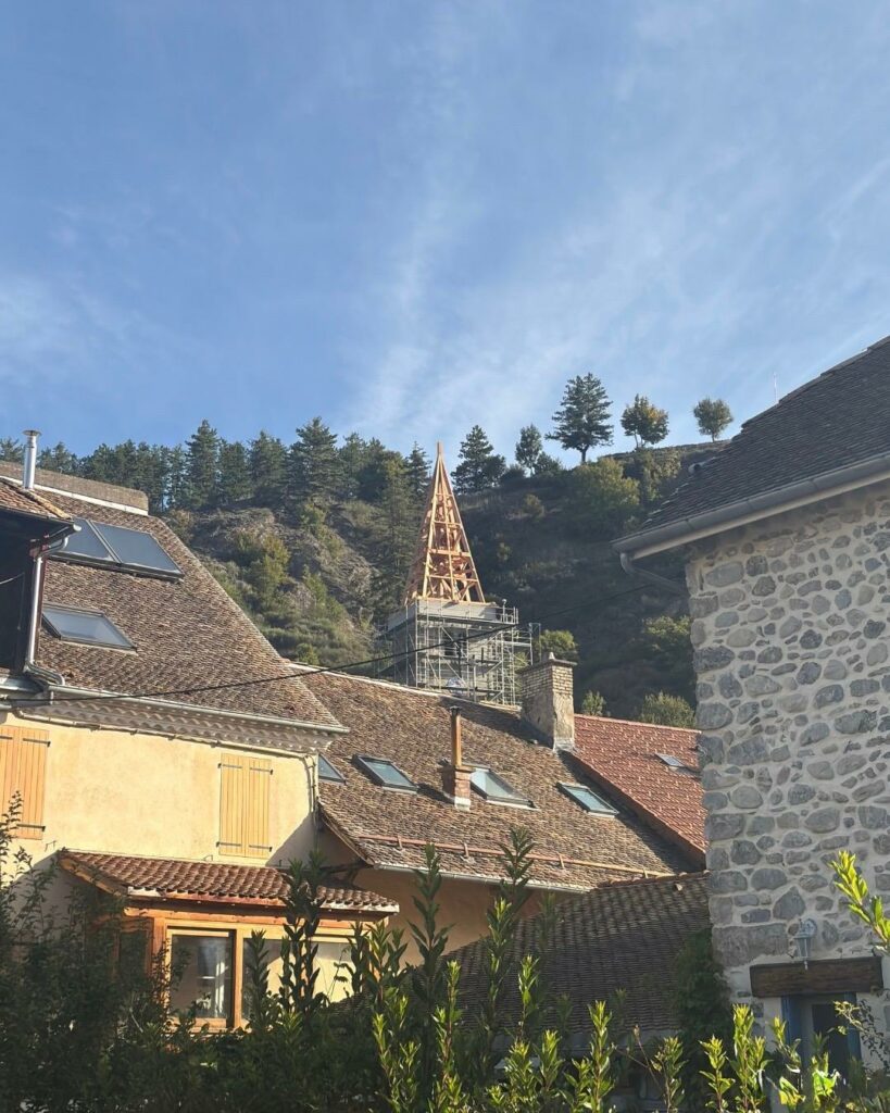 Vue sur les toits en tuiles d’un village, avec en arrière-plan le clocher d’une église en restauration entouré d’échafaudages, au pied d’une colline boisée sous un ciel bleu