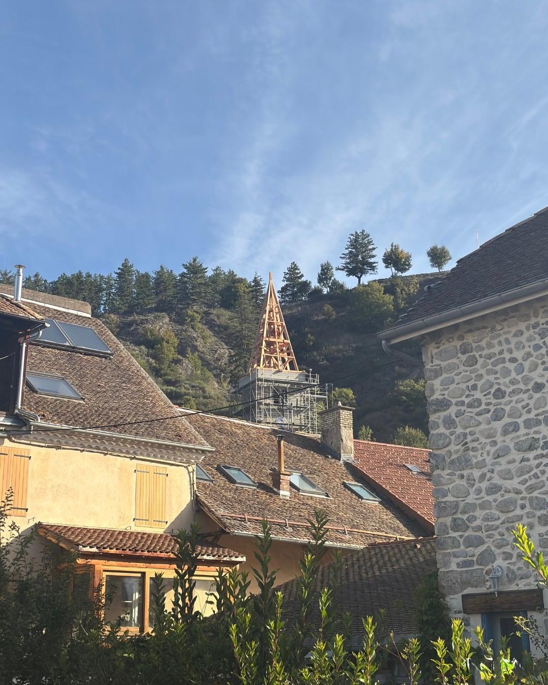 Vue sur les toits en tuiles d’un village, avec en arrière-plan le clocher d’une église en restauration entouré d’échafaudages, au pied d’une colline boisée sous un ciel bleu