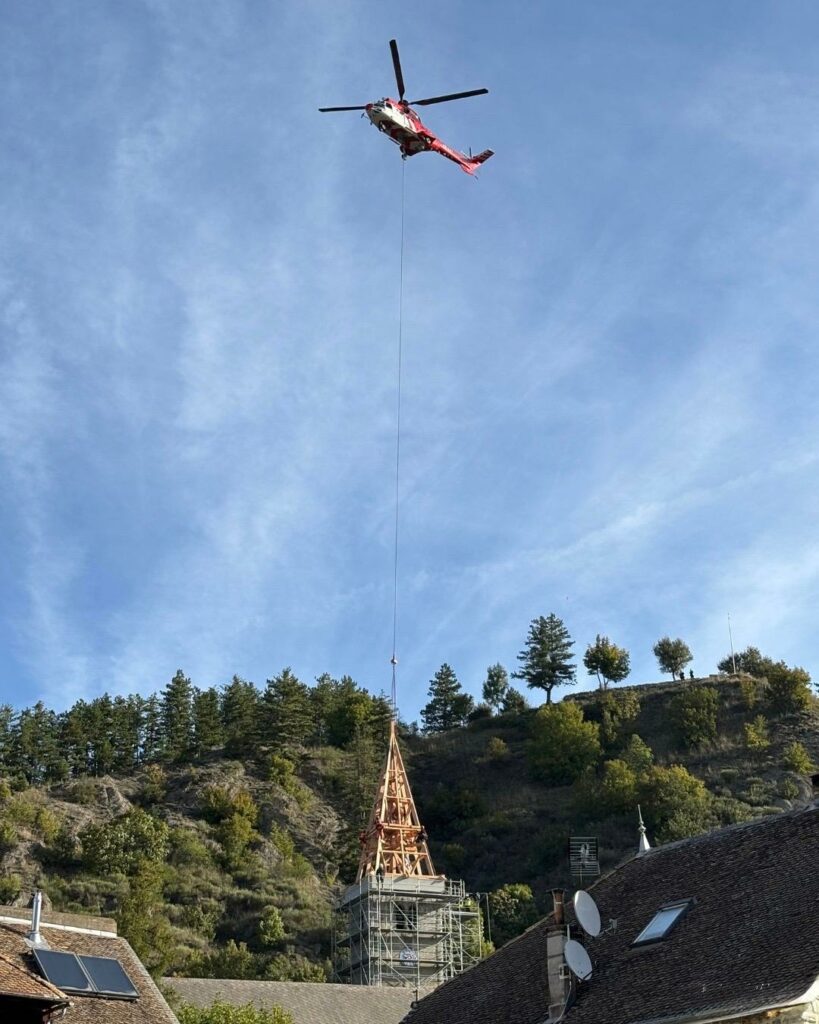 Hélicoptère rouge transportant par câble la flèche en bois d’un clocher en restauration, au-dessus d’un village et d’une colline boisée sous un ciel bleu.
