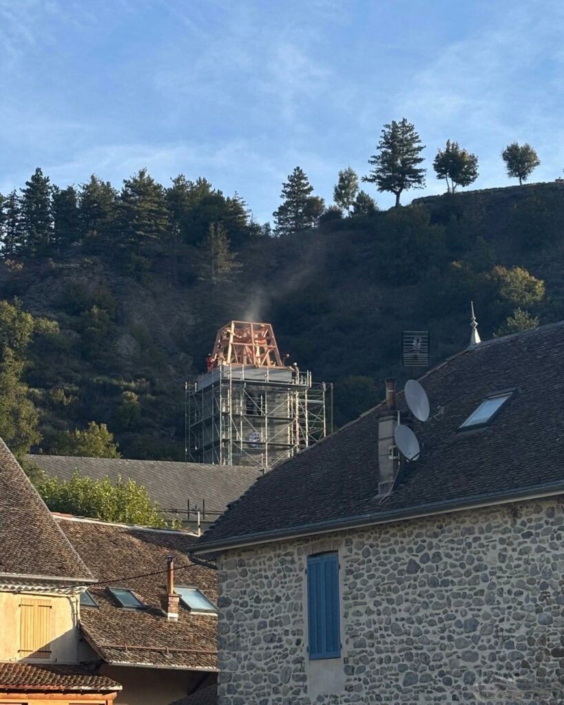 Vue sur les toits en tuiles d’un village, avec en arrière-plan le clocher d’une église en restauration entouré d’échafaudages, au pied d’une colline boisée sous un ciel bleu