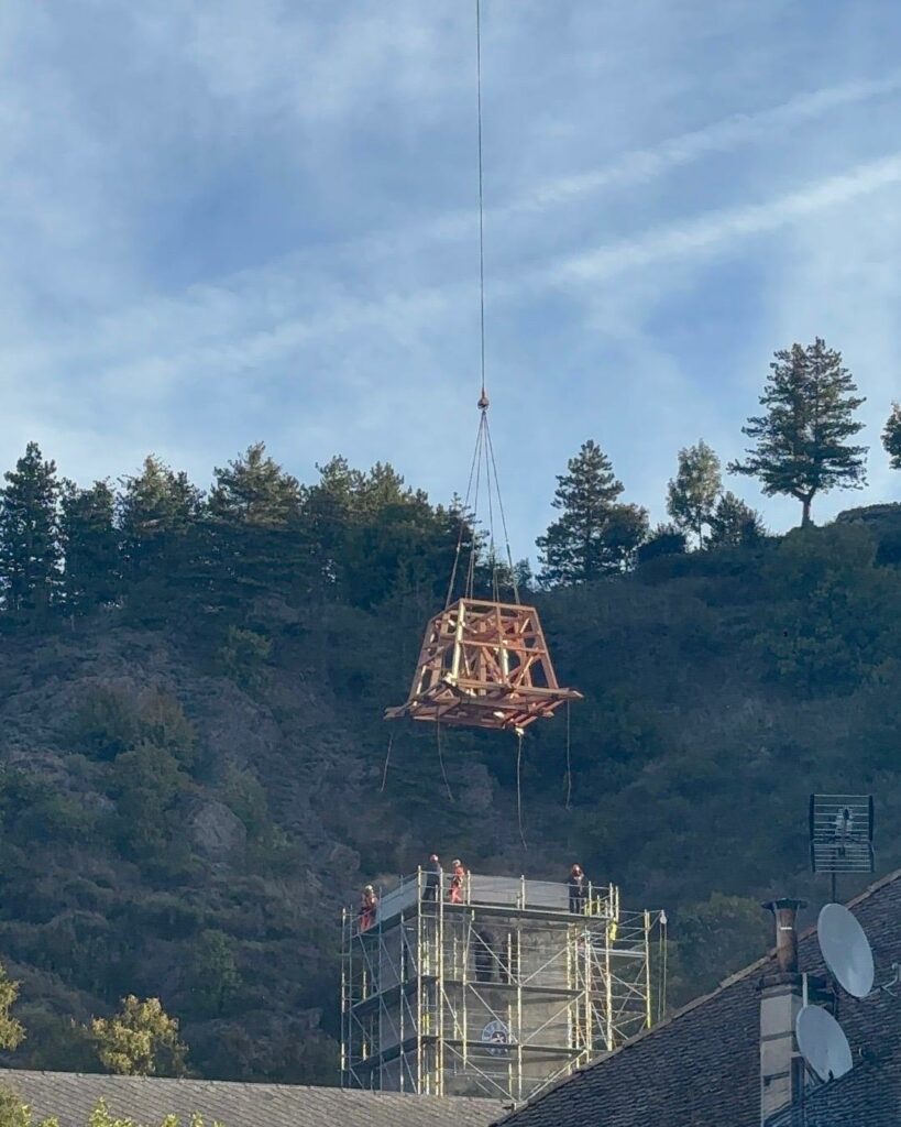 Vue sur les toits en tuiles d’un village, avec en arrière-plan le clocher d’une église en restauration entouré d’échafaudages, au pied d’une colline boisée sous un ciel bleu.”