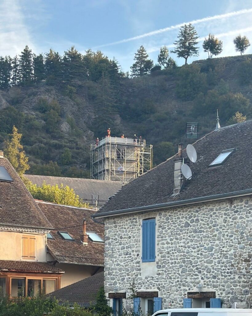 Vue sur les toits en tuiles d’un village, avec en arrière-plan le clocher d’une église en restauration entouré d’échafaudages, au pied d’une colline boisée sous un ciel bleu.”