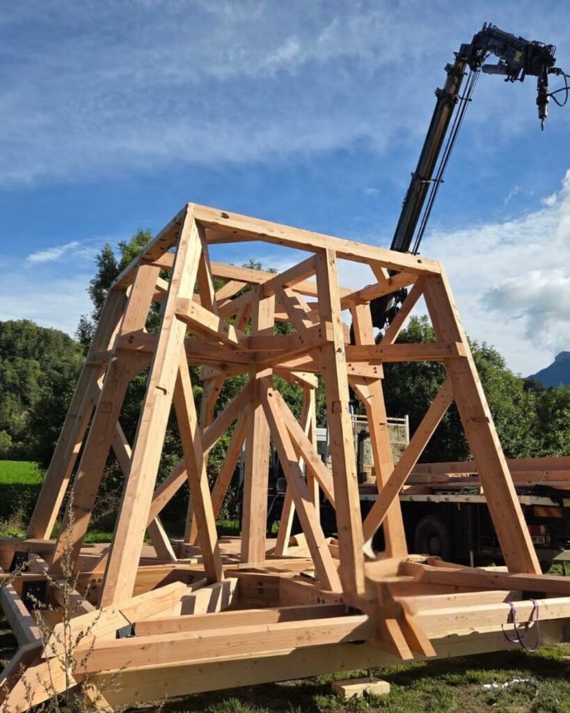 Structure en bois d’un clocher en cours d’assemblage sur un chantier, avec une grue mobile à l’arrière-plan sous un ciel bleu.