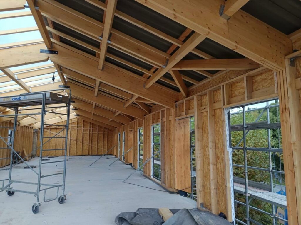 Intérieur d’un bâtiment en ossature bois en cours de construction, montrant la charpente apparente, les ouvertures murales et un échafaudage mobile sur un sol encore brut.