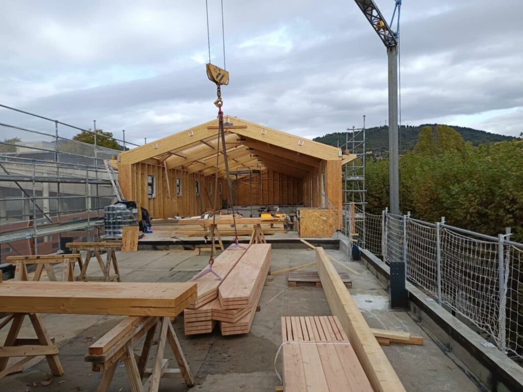 Chantier de construction d’un bâtiment en ossature bois, avec une grue soulevant des poutres et l’intérieur de la structure encore ouvert, entouré d’échafaudages sous un ciel nuageux.